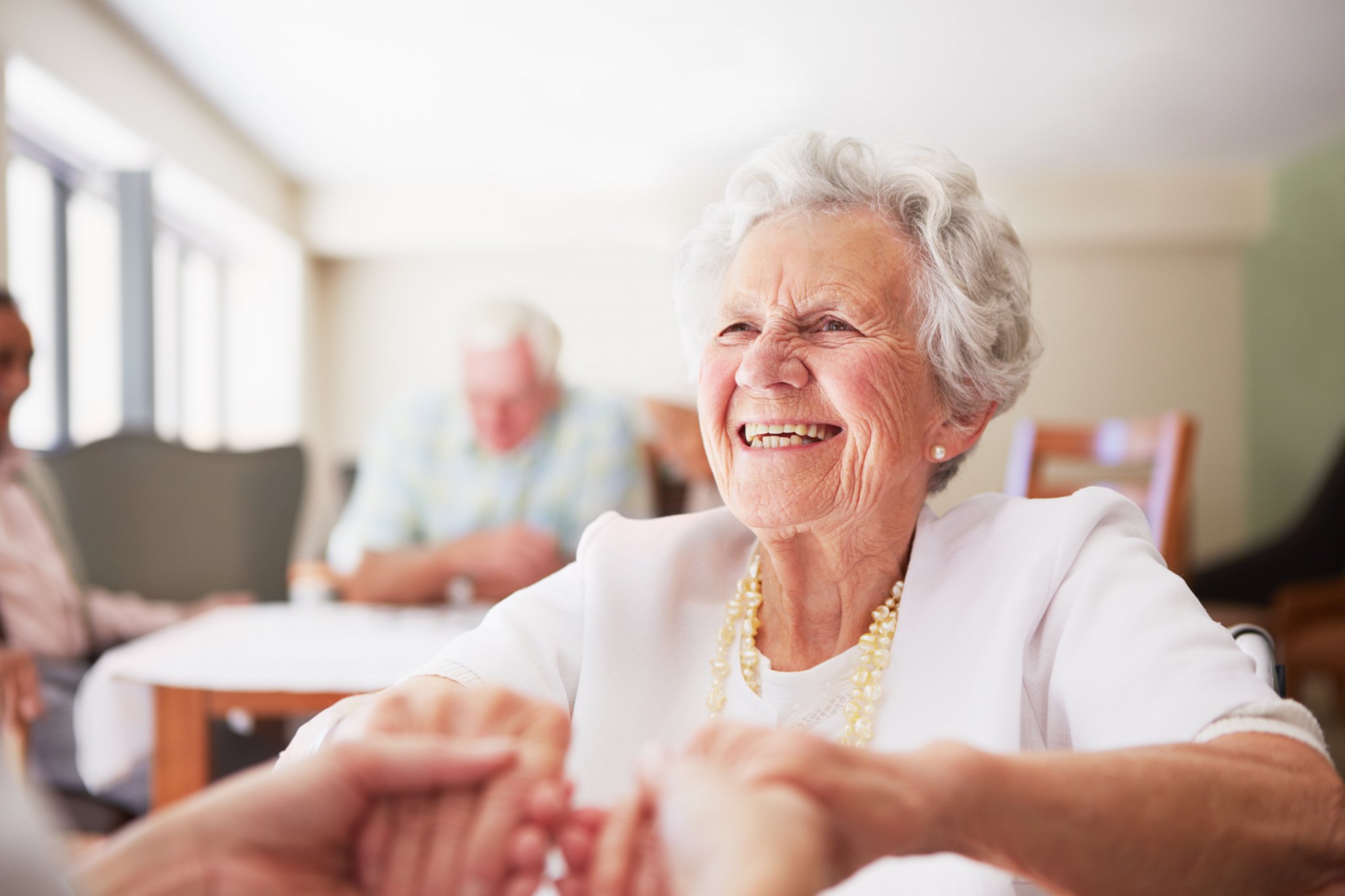 Elderly woman smiling while holding hands in a bright communal room.